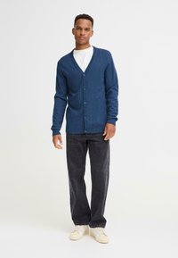 Cardigan blu navy con scollatura a V, chiusura con bottoni e maniche lunghe, abbinato a jeans scuri e sneaker chiare. Texture liscia.