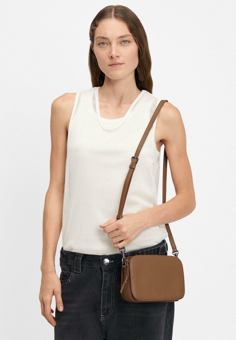 Borsa a tracolla in pelle marrone con forma rettangolare, tracolla regolabile e branding discreto. Indossata con un top senza maniche bianco e jeans scuri.