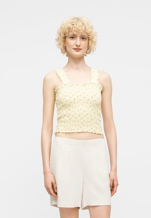 Jeune personne aux cheveux blonds bouclés et courts, portant un débardeur jaune à smocks fleuri et un short blanc taille haute, debout devant un fond blanc.