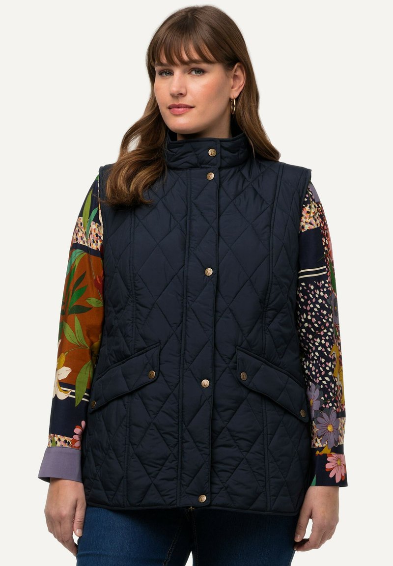 Marineblaues, gestepptes Weste mit hohem Kragen, ausgestattet mit zwei Fronttaschen und goldenen Druckknöpfen. Dazu wird ein florales Langarmshirt darunter getragen.