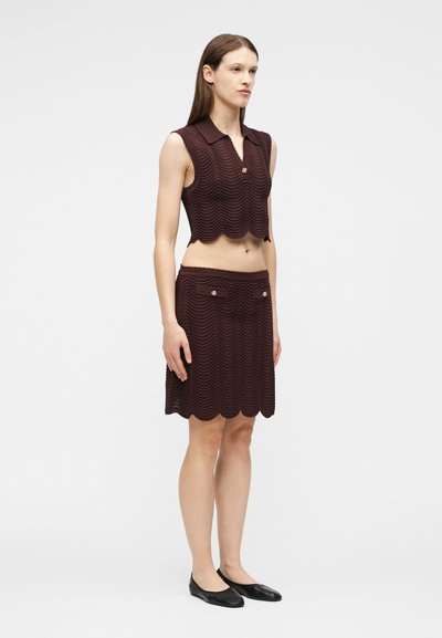 Femme debout portant un crop top texturé sans manches marron foncé avec col et une jupe assortie mi-longue avec poches avant et des ballerines noires.