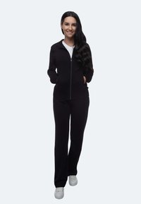 Schwarze Zip-Jacke mit Kragen und Taschen, kombiniert mit passenden Hosen. Das Model trägt ein weißes Hemd darunter und vervollständigt den Look mit weißen Schuhen.