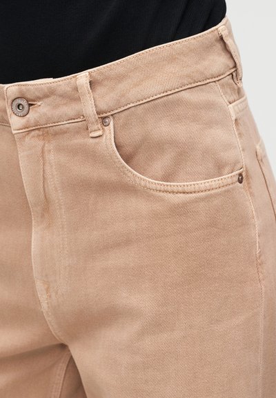 Κοντινό πλάνο στη μέση και στην μπροστινή τσέπη μπεζ παντελονιού από τζιν, με ορατό κουμπί και θηλιά για ζώνη, φορεμένο με μαύρο επάνω μέρος.