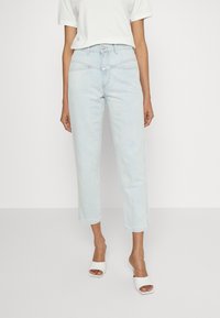 Kvinna som bär ljusblå högmidjade avklippta jeans, en vitkortärmad topp och vita öppna klackskor, stående mot en enfärgad bakgrund.