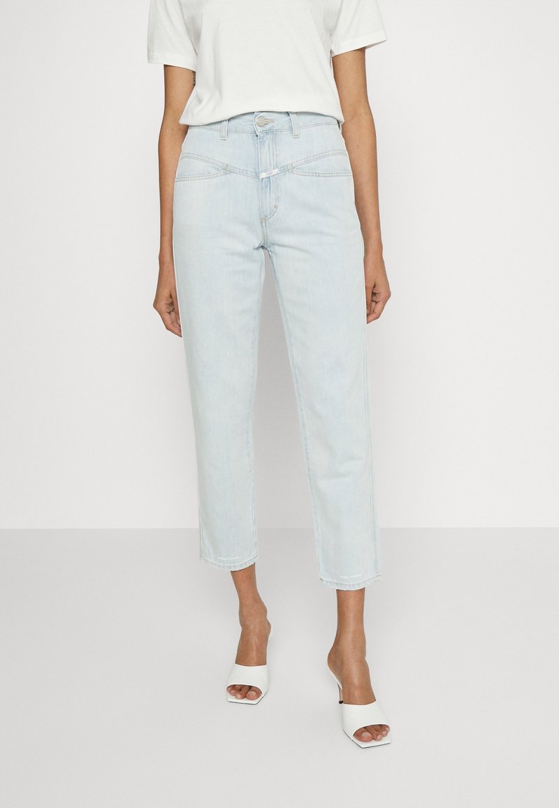 Kvinna som bär ljusblå högmidjade avklippta jeans, en vitkortärmad topp och vita öppna klackskor, stående mot en enfärgad bakgrund.