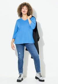 Blaues, V-Ausschnitt, langärmliges Oberteil kombiniert mit ausgewaschen blauen Jeans und silbernen Plateau-Stiefeln, mit einem schwarzen Pullover, der über dem Arm hängt.