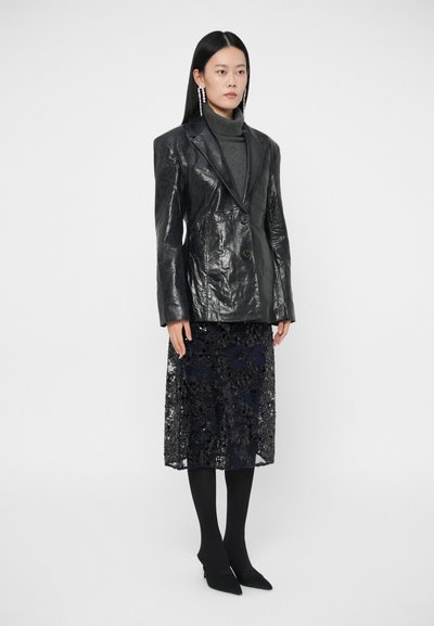 Czarny skórzany żakiet z wyraźnie zaznaczonymi ramionami i dwoma guzikami, zestawiony z przezroczystą czarną haftowaną spódnicą na ciemnych legginsach i spiczastych butach.