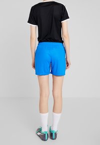 Svart träningstop med kort ärm och vita detaljer, kombinerad med blå träningsshorts. Synliga sportskor och vita strumpor. Enkelt bakgrund.