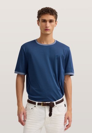 Junger Mann mit kurzen lockigen Haaren, der ein blaues kurzärmliges Hemd in weiße Hose gesteckt trägt und einen geflochtenen braunen Gürtel trägt.