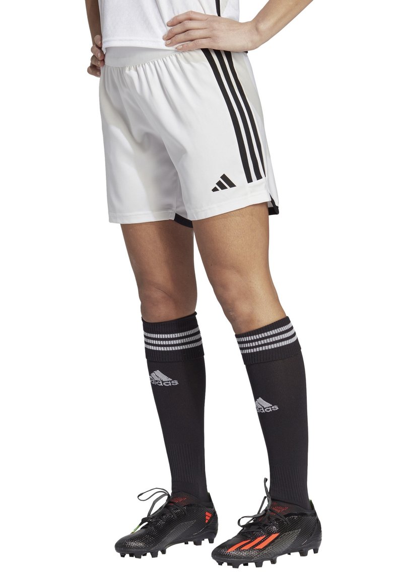 Weiße Sportshorts mit schwarzem Dreistreifen-Design, kombiniert mit schwarzen Socken mit weißen Streifen und schwarzen Fußballschuhen mit roten Akzenten.