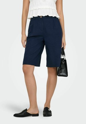 Vrouw draagt marineblauwe op maat gemaakte knielange shorts, witte mouwloze top met ruches, zwarte instap loafers, houdt een zwarte textuur handtas vast.