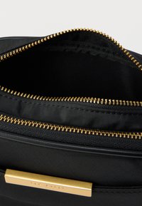 Μαύρη υφασμάτινη τσάντα με χρυστό φερμουάρ και μεταλλικά εξαρτήματα. Το εσωτερικό διαθέτει υφή επένδυσης και πλακέτα με το όνομα της μάρκας. Ανοιχτό πάνω μέρος που δείχνει τις λεπτομέρειες του χώρου αποθήκευσης.