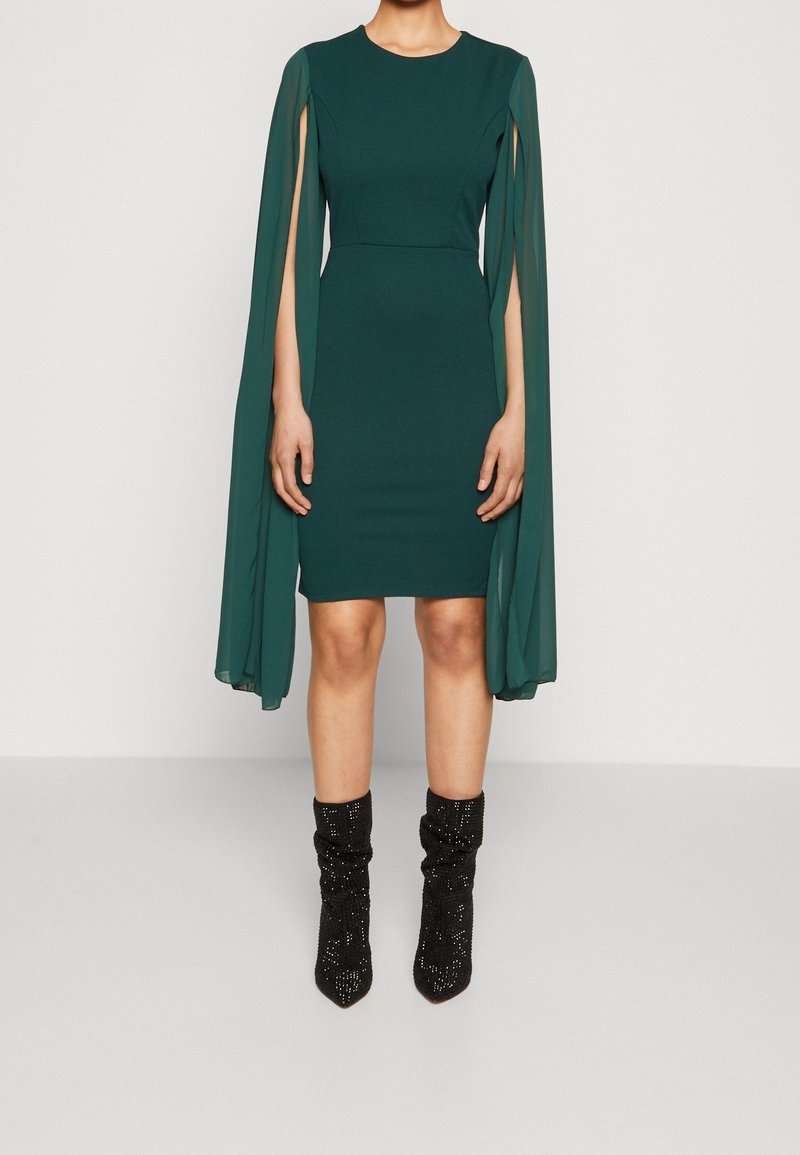 Femme portant une robe vert foncé longueur genou avec des manches longues ouvertes style cape et des bottines noires ornées de strass.