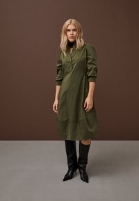 Plavuša u midi haljini maslinasto zelene boje s dugim rukavima i crnim čizmama do koljena sa špicastim vrhom, stoji na smeđoj i sivoj pozadini.