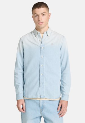 Młody mężczyzna ubrany w jasnoniebieską, zapiętą na guziki koszulę z długim rękawem oraz dopasowane dżinsy, stojący na tle jednolitej białej ściany.