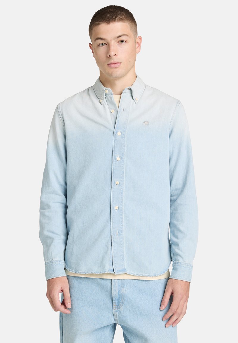 Jeune homme portant une chemise à manches longues boutonnée bleu clair et un jean assorti, debout devant un fond blanc uni.