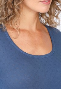 Blue top made of textured fabric with small patterns, featuring a rounded neckline and scalloped edging. Soft and fitted design.