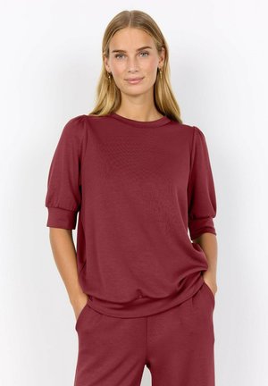 Vrouw met stijl blond haar die een kastanjebruine top met pofmouwen en bijpassende broek draagt, handen in haar zakken, staand tegen een witte achtergrond.