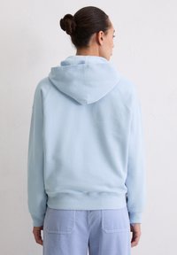 Kobieta z kokiem na głowie, ubrana w jasnoniebieską bluzę z kapturem i niebieskie spodnie w paski, stojąca tyłem do białej, strukturalnej ściany.