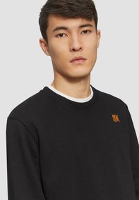 Sweatshirt noir avec un col rond, arborant un patch logo orange sur la poitrine. Le tissu semble doux avec une texture lisse.