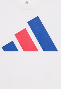 Bílá bavlněná tričko s velkým potiskem, zahrnujícím tři šikmé tvary v červené a modré barvě, připomínající logo značky. Hladká textilní struktura.