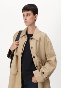 Beige overskjorte laget av bomull, med en stor krage, svarte knapper og subtile lomme-detaljer. Båret over en svart sweatshirt med tekstur.