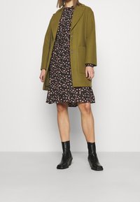 Olivgrön ullkappa med två knappar och sidofickor över en svart blommig klänning. Svarta ankelskór fulländar outfiten.