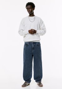 Felpa grigia, jeans blu larghi e sneakers grigio-marroni. Dettagli notevoli includono collane sovrapposte e una vestibilità comoda.