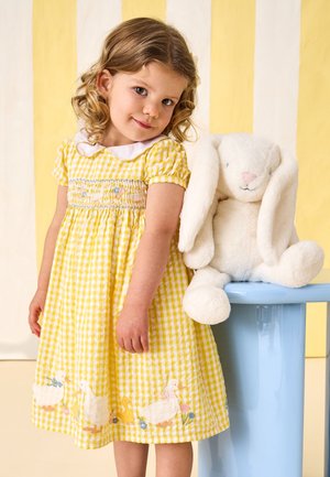 Fiatal lány sárga kockás ruhában, amelyen kacsa hímzés van, egy nagy fehér plüssnyúl mellett áll egy kék széken.