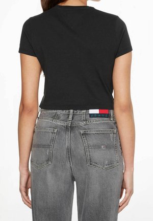Femme portant un T-shirt court noir et un jean gris taille haute avec poches arrière et une petite étiquette rouge, blanche et bleue.