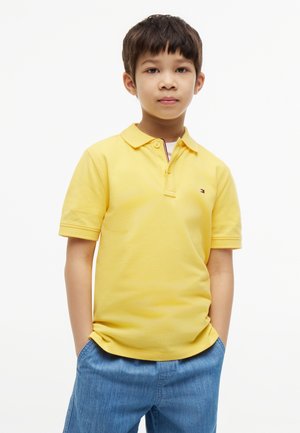Junge trägt ein gelbes kurzärmeliges Poloshirt und blaue Jeans mit den Händen in den Taschen, steht vor einem weißen Hintergrund.
