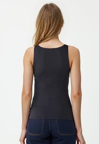 Czarny top bez rękawów o gładkiej teksturze, zaokrąglonym dekolcie i dopasowanym kroju. Noszony z ciemnoniebieskimi dżinsami.