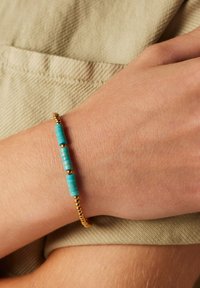 Bracelet en perles dorées et turquoise avec des perles cylindriques en turquoise et des perles rondes en or. Porté sur un poignet, sur un fond en tissu beige texturé.