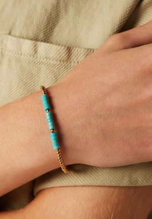 Gold and turquoise bead bracelet with cylindrical turquoise and round gold beads. Worn on a wrist against a beige textured fabric background.
