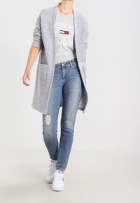 Cardigan largo gris claro hecho de una tela suave con un corte relajado, combinado con una camiseta gris con texto, jeans azul desgastados y zapatillas blancas.