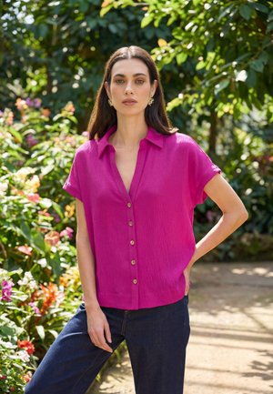 Donna in piedi all'aperto con la mano sul fianco, indossa una camicia magenta a maniche corte con bottoni e jeans blu scuro, circondata da vegetazione e fiori.