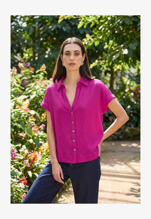 Donna in piedi all'aperto con la mano sul fianco, indossa una camicia magenta a maniche corte con bottoni e jeans blu scuro, circondata da vegetazione e fiori.
