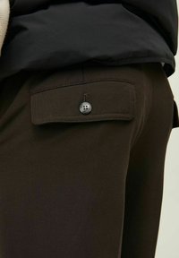 Pantalones marrones con un bolsillo trasero con solapa y cierre de botón único. La tela parece suave con un acabado mate.