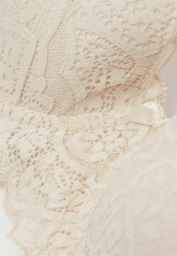 Kanten beha in zacht beige met ingewikkelde bloemmotieven; heeft een gestructureerd oppervlak en een delicate lintaccent bij de band.