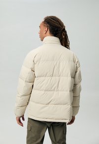 Man met dreadlocks in een beige pufferjack en olijfgroene cargobroek, staand met zijn rug naar de camera tegen een lichtgrijze achtergrond.