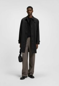 Black long coat with a curved collar, button closure, relaxed fit, worn over a dark top and light brown wide-leg trousers. Holds a small bag.