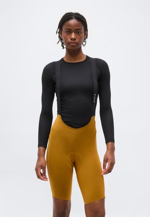 Femme portant un haut noir à manches longues ajusté et un cuissard de cyclisme moutarde avec bretelles, debout devant un fond blanc uni.