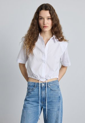 Junge Frau mit langen lockigen Haaren, die ein weißes kurz geschnittenes Hemd mit Knöpfen und hoch taillierte blaue Jeans trägt, vor einem einfarbigen Hintergrund.