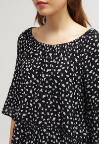 Schwarze Bluse mit weißen Blumenmustern, weitem Ausschnitt, gerüschter Textur und kurzen Ärmeln. Der Stoff wirkt leicht und leicht zerknittert.