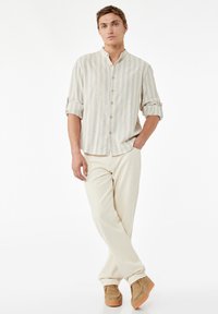 Herre skjorte med knapper i lys beige og hvide striber, med en ståkrave og optrukne ærmer. Parret med cremede bukser og brune sko.