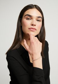 Sudraba aproce ar caurspīdīgiem dārgakmeņiem, eleganti aizdarīta. Modelis ģērbjas melnā blūzē, roka domīgi novietota zem zoda. Gluda, spīdīga virsma.