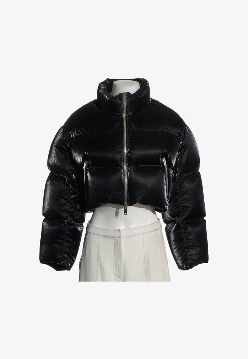 Veste courte matelassée noire brillante avec col montant, fermeture éclair à l'avant et poches zippées, présentée sur un mannequin blanc portant un pantalon clair.