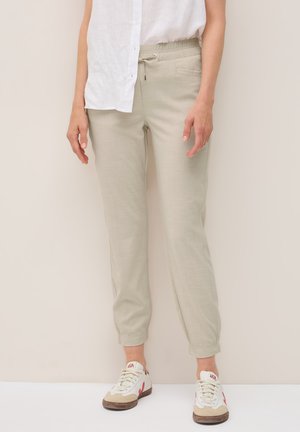 Una mujer está de pie, vistiendo una camisa blanca y pantalones jogger beige claros, combinados con zapatillas casuales, frente a un fondo neutro.