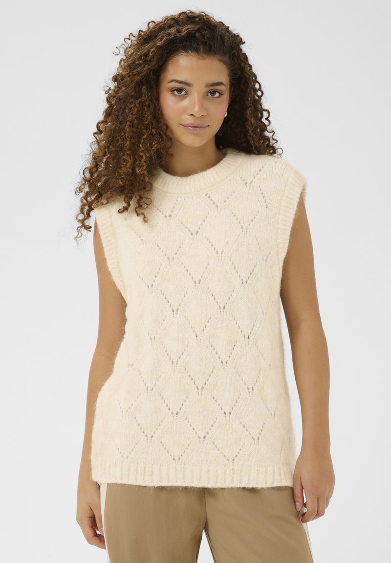 Žena s kovrčavom kosom koja nosi bezrukavu kremastu pletenu vestu s dijamantnim uzorkom i smeđe hlače, na jednobojnoj pozadini.