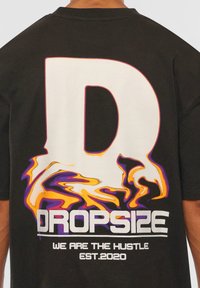 Musta t-paita, jossa on suuri, tyylitelty valkoinen "D" värikkäillä liekkimäisillä grafiikoilla ja rohkealla tekstillä: "DROPSIZE ME OLEMME HUSTLE EST.2020."